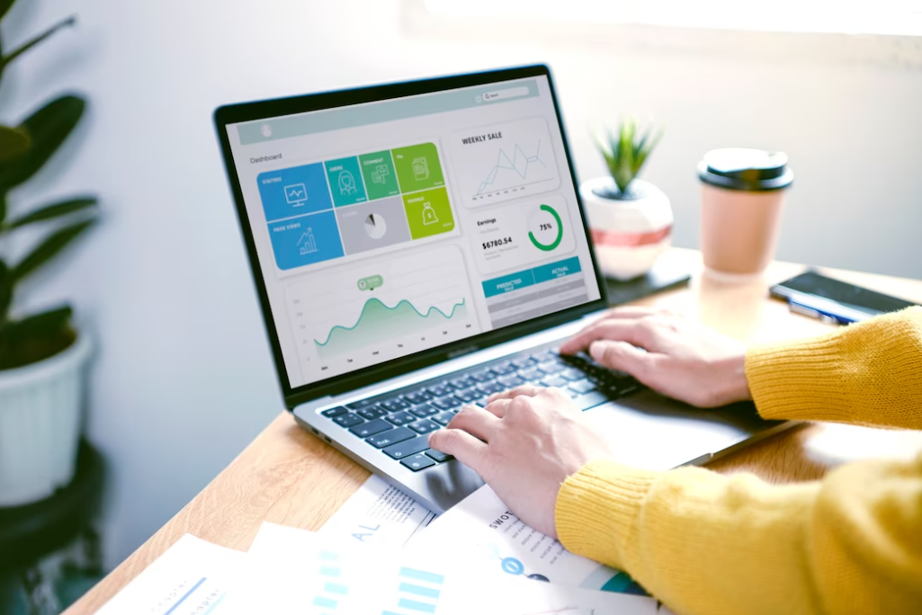 person in a yellow sweater typing on the laptop keyboard with analytics on it, a cup of coffee, and a vase with a flower in the corner of the table