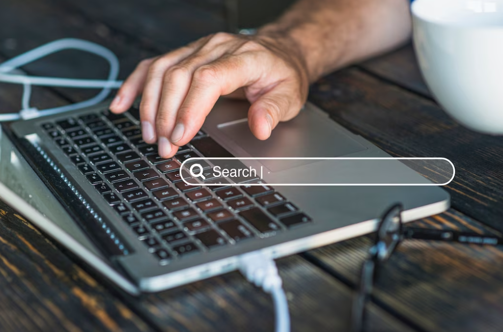 hand on a laptop keyboard, glasses, and a white cup on  the wooden table